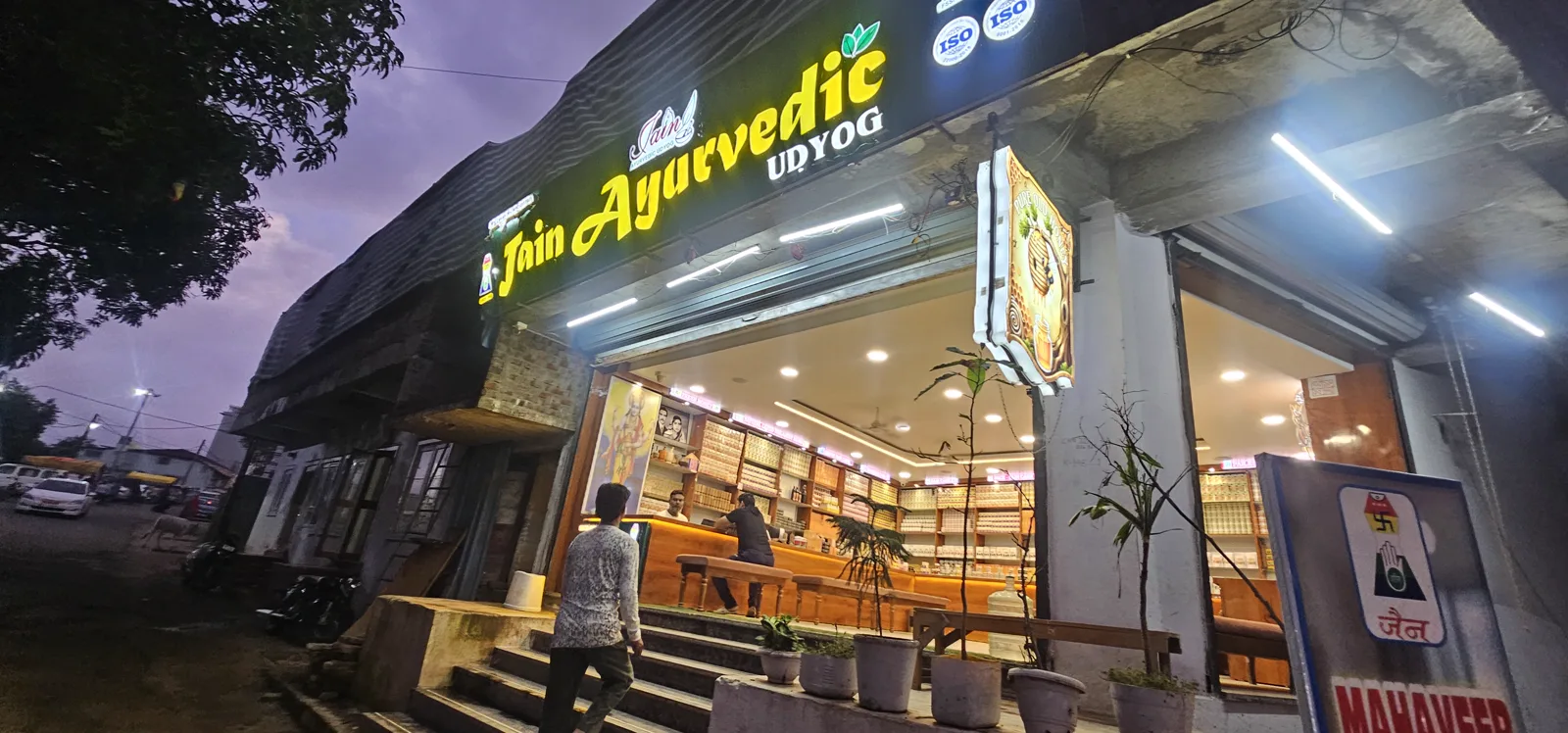 The lit storefront of Jain Ayurvedic Udyog, a herbal and honey shop in the Pachmarhi market, at dusk