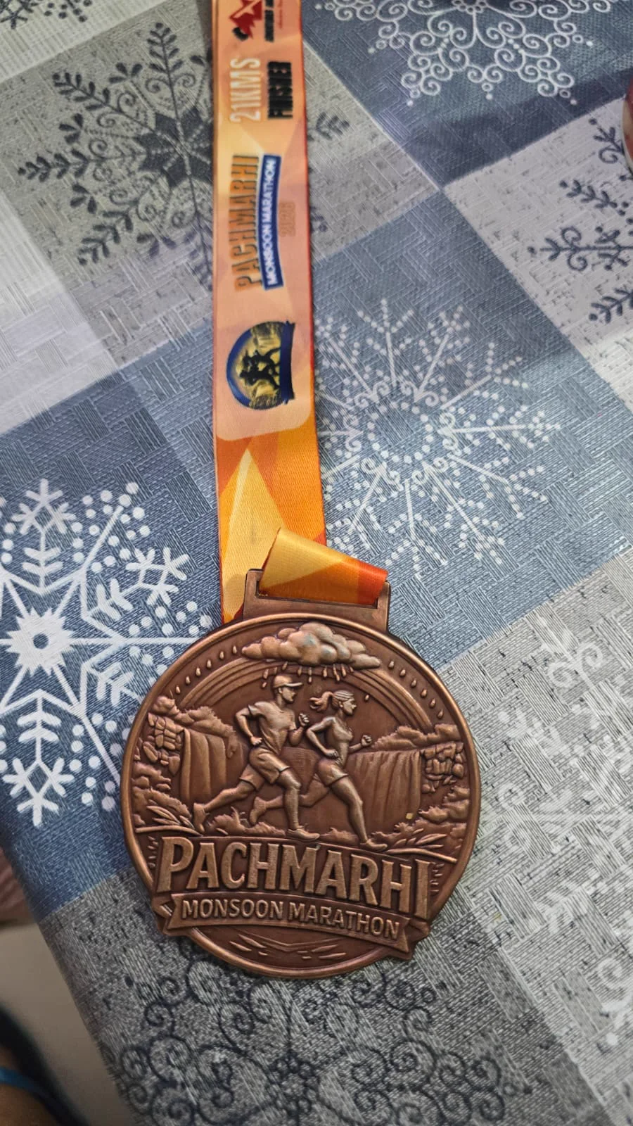 Close-up of the Pachmarhi Monsoon Marathon 2026 finisher medal for 21 km, showing rain clouds, two runners and a waterfall in relief